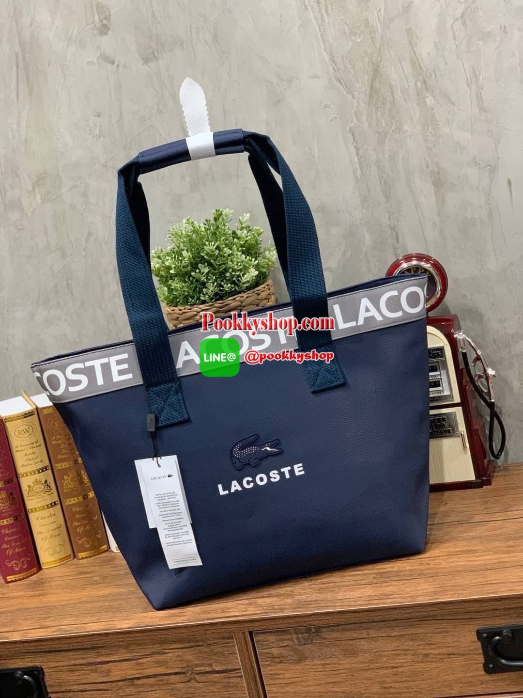 ลาคอส Shopping Bag กระเป๋าถือทรงช้อปปิ้งขนาดใหญ่ ที่ใส่สัมภาระได้รวมถึงใส่เอกสารขนาด A4 ได้ วัสดุ PVC กันน้ำ ดูแลรักษาง่าย ตัดขอบด้วยแถบโลโก้สีเทา ด้านหน้าสกรีนโลโก้และประดับจรเข้อันเป็นสัญลักษณ์ เปิดปิดด้วยซิปเดียว ภายในสกรีนโลโก้แบรนด์ ใบใหญ่สวยโดดเด่