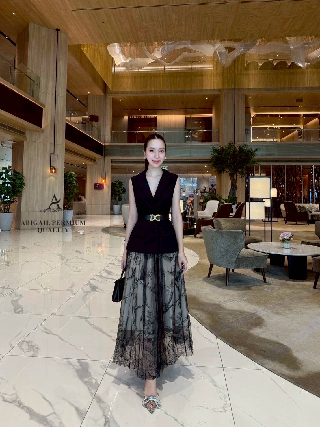 เสื้อแขนกุดเบลเซอร์สีดำ ดีไซน์คอวีลึก เพิ่มความเรียบหรูพร้อมกระดุมด้านหน้าและกระเป๋าดีไซน์เรียบเท่ ดึงความสนใจด้วยเข็มขัดหนังหัวสีทองที่ช่วยเน้นทรวดทรงและเพิ่มความพรีเมียม กระโปรงยาวทรงเอ ผ้าซีทรูสองชั้น ด้านในเป็นผ้าสีเบจเรียบ ส่วนด้านนอกเป็นผ้าทูลล์สีดำ