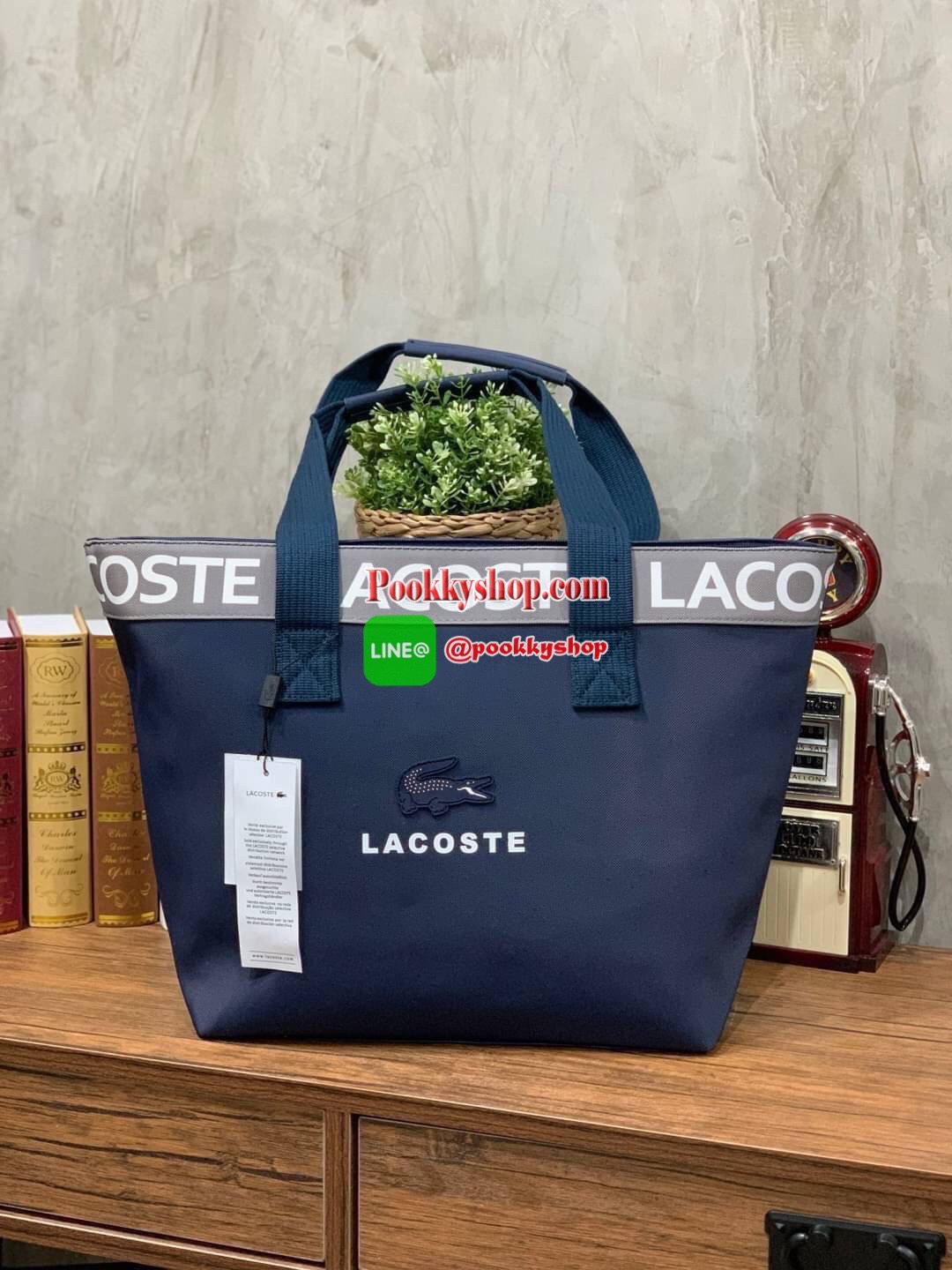 ลาคอส Shopping Bag กระเป๋าถือทรงช้อปปิ้งขนาดใหญ่ ที่ใส่สัมภาระได้รวมถึงใส่เอกสารขนาด A4 ได้ วัสดุ PVC กันน้ำ ดูแลรักษาง่าย ตัดขอบด้วยแถบโลโก้สีเทา ด้านหน้าสกรีนโลโก้และประดับจรเข้อันเป็นสัญลักษณ์ เปิดปิดด้วยซิปเดียว ภายในสกรีนโลโก้แบรนด์ ใบใหญ่สวยโดดเด่